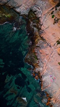 Drone video showing rugged Spanish coastline with turquoise sea, clear shallow waters, and small groups of people relaxing on the rocks.