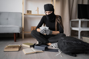Female thief in black balaclava counting stolen money from house sitting on floor with cash