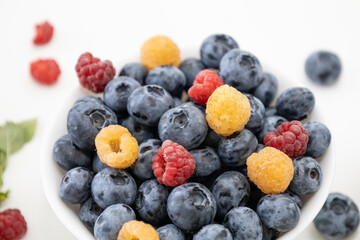 Blueberry and raspberry is tasty, healthy and delicious fruits. Fresh blueberries with red and yellow raspberries with leaves of mint in white bowl. Wild berry in white plate on white background.