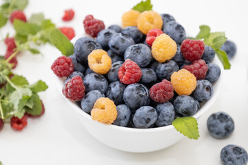 Blueberry and raspberry is tasty, healthy and delicious fruits. Fresh blueberries with red and yellow raspberries with leaves of mint in white bowl. Wild berry in white plate on white background.