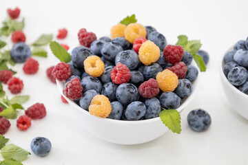 Blueberry and raspberry is tasty, healthy and delicious fruits. Fresh blueberries with red and yellow raspberries with leaves of mint in white bowl. Wild berry in white plate on white background.