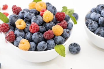Blueberry and raspberry is tasty, healthy and delicious fruits. Fresh blueberries with red and yellow raspberries with leaves of mint in white bowl. Wild berry in white plate on white background.