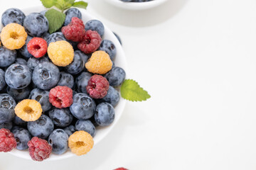 Blueberry and raspberry is tasty, healthy and delicious fruits. Fresh blueberries with red and yellow raspberries with leaves of mint in white bowl. Wild berry in white plate on white background.
