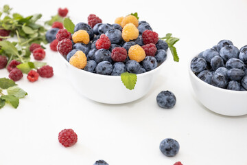 Blueberry and raspberry is tasty, healthy and delicious fruits. Fresh blueberries with red and yellow raspberries with leaves of mint in white bowl. Wild berry in white plate on white background.