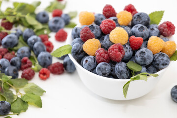 Blueberry and raspberry is tasty, healthy and delicious fruits. Fresh blueberries with red and yellow raspberries with leaves of mint in white bowl. Wild berry in white plate on white background.