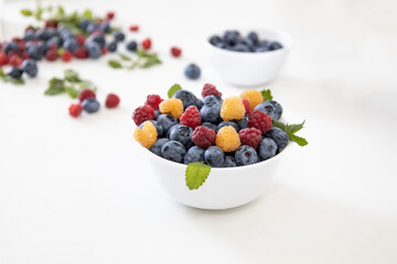 Blueberry and raspberry is tasty, healthy and delicious fruits. Fresh blueberries with red and yellow raspberries with leaves of mint in white bowl. Wild berry in white plate on white background.