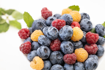 Blueberry and raspberry is tasty, healthy and delicious fruits. Fresh blueberries with red and yellow raspberries with leaves of mint in white bowl. Wild berry in white plate on white background.