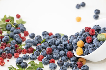 Blueberry and raspberry is tasty, healthy and delicious fruits. Fresh blueberries with red and yellow raspberries with leaves of mint in white bowl. Wild berry in white plate on white background.