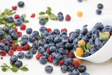 Blueberry and raspberry is tasty, healthy and delicious fruits. Fresh blueberries with red and yellow raspberries with leaves of mint in white bowl. Wild berry in white plate on white background.