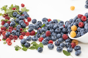 Blueberry and raspberry is tasty, healthy and delicious fruits. Fresh blueberries with red and yellow raspberries with leaves of mint in white bowl. Wild berry in white plate on white background.