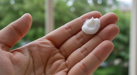 A hand holding a dollop of white cream Green foliage background