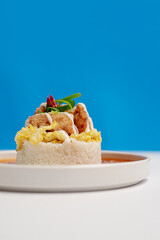 A serving of rice topped with soft scrambled eggs, crispy karaage chicken, special savory sauce, and fresh chili and scallion garnish on a minimalist white plate with a modern white & blue background.