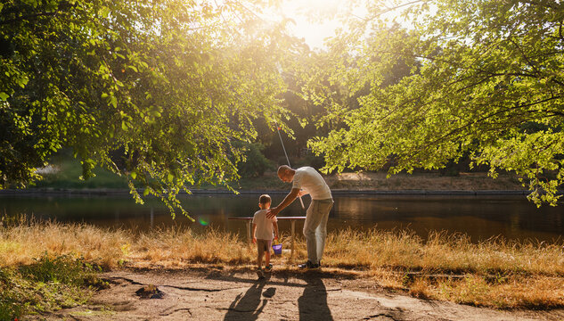 Father and young son fishing at calm lake in countryside at sunset. Horizontal family lifestyle photo with nature background and warm summer mood.
