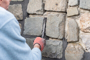 Builder applies mortar between stones in a wall using a long, narrow trowel during renovation work,...