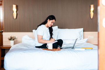 An Asian woman sitting cross-legged on a bed, writing in a notebook with a laptop and coffee mug nearby, representing remote work, planning, and creative thinking in a relaxed bedroom environment.