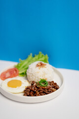 Close-up of cumin-spiced beef served with steamed white rice, sunny side up egg, fresh lettuce, and tomato slices on a white plate, photographed on a clean minimal background.
