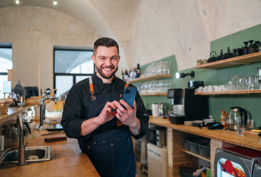 Smiling small business owner dressed in a black chef uniform with an apron using smartphone in his own cozy restaurant hall. Successful people, hard work, consumer cafes restaurants industry concept - Powered by Adobe