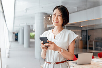 Asian mature business woman manager ceo using cell phone mobile app. Smiling eastern 40s years old female businesswoman holding smartphone in office workspace for trading, banking, working online