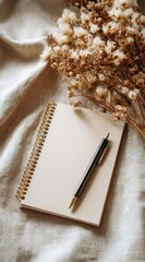 Spiral notebook and pen on linen fabric with dried flowers create a cozy and artistic workspace setting.