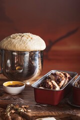 Dough rises in a silver bowl beside chocolate-filled babka dough in bread pans, ready for baking. Walnuts and butter rest nearby on the rustic wooden surface