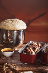 A metal bowl overflows with risen dough near baking tins filled with sweet swirls. Walnuts and butter sit nearby, promising a delicious babka on a rustic wooden surface