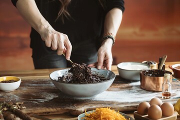 A person mixes a dark, rich batter in a bowl, likely for a Christmas cake. Eggs, shredded fruit, nuts, and other ingredients are scattered around on a wooden surface, ready to be added