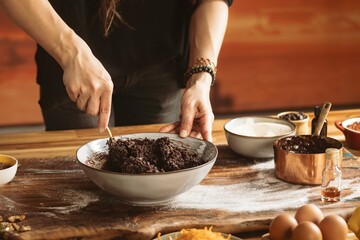 Hands mix a dark brownie batter in a grey bowl on a wooden table. Ingredients like eggs, sugar, oil, and cocoa are spread around her, ready to be incorporated