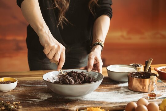 A baker carefully stirs a dark, rich mixture in a bowl on a floured wooden surface. Various ingredients in containers and eggs are scattered around, preparing for the next step - Powered by Adobe
