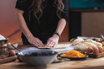 Against a warm kitchen backdrop, a woman kneads dough on a wooden surface dusted with flour. Bowls with fillings and eggs sit nearby, ready for preparing traditional Eastern European bread
