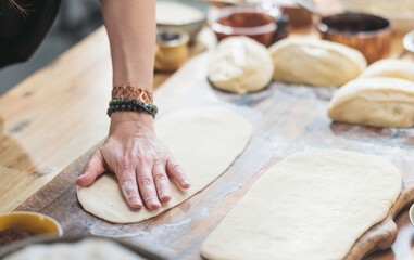 A woman's hands press and shape a pale dough on a wooden board atop a kitchen island, with several portions of dough and toppings around, preparing it for pizza night