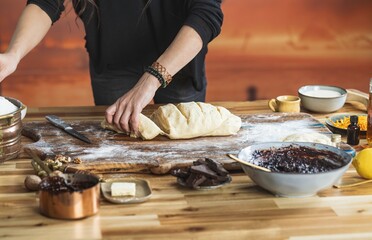 A person with bracelets prepares dough on a flour-dusted wooden board. Ingredients such as jam, chocolate, butter, and sugar are nearby, ready for baking holiday goodies