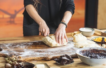 On a wooden table dusted with flour, a person uses a knife to cut dough. Bowls of chocolate, jam, and other baking ingredients surround the kneading surface