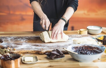 A person is cutting a large dough ball on a wooden cutting board, surrounded by ingredients like chocolate, jam, and butter, in a cozy kitchen bathed in warm light