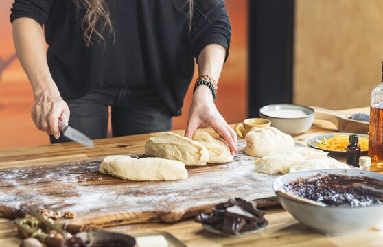 A baker expertly divides soft dough with a knife on a flour-dusted wooden surface. Sweet fillings, butter, and other baking ingredients are scattered around the table - Powered by Adobe