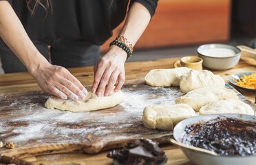 A person kneads and shapes dough on a flour-dusted wooden surface, preparing for baking. Other ingredients and tools are visible, suggesting a baking process is in progress