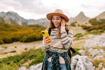 Beautiful woman in cozy sweater and hat uses smartphone against backdrop of mountain landscape. Young woman hiker with a phone on trail in mountains. Concept of nature, freedom. Active lifestyle.