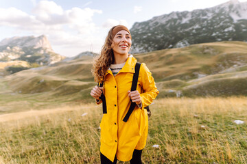 Cheerful woman in bright raincoat explores mountainous terrain at sunset. Young female traveler with backpack feels freedom and enjoys mountain landscape. Nature, adventure concept.
