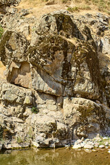 Rock face with moss and lichens by the water’s edge of a mountain stream in Boshkizilsay, Uzbekistan. Sunny summer day, view of the natural shore and water reflection