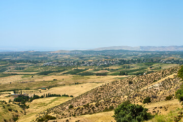 Naklejka premium A panoramic view of the rural landscape in the foothills of Uzbekistan. Yellow, arid hills contrast with green fields and vineyards, with a small settlement visible in the distance. July 2025.