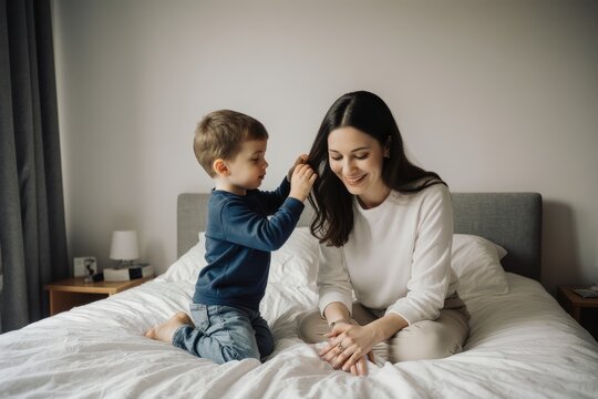 Cute son combing mother's hair while kneeling on bed against wall at home