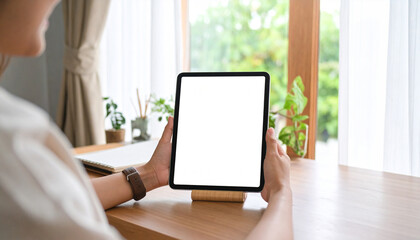 Fototapeta premium Over the shoulder view of a woman holding a digital tablet with a blank screen mockup, sitting at a desk in a cozy home office.