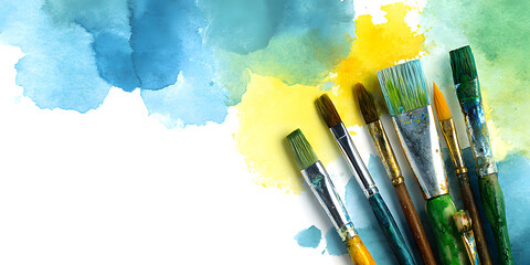 Obraz premium Colorful watercolor splashes in blue, yellow, and green frame paintbrushes and paints on a clean white background for creative stationery use.