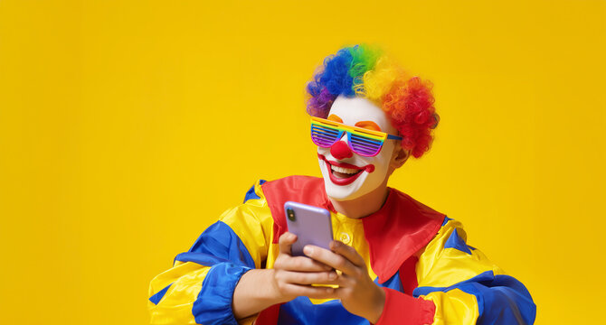 Clown in colorful costume with rainbow wig and shutter shades using smartphone on yellow background.