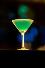 Green cocktail garnished with lime, resting on wooden bar surface, blurred lights highlighting elegant mixology scene