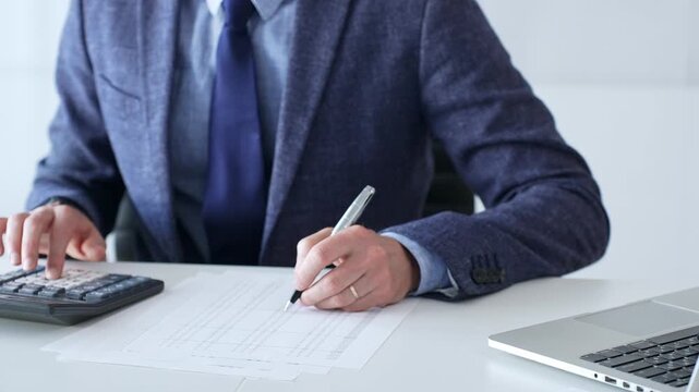Businessman wearing suit and tie is using calculator and taking notes while working at desk in office, with laptop and stack of folders nearby, close up. Audit and taxes theme in business