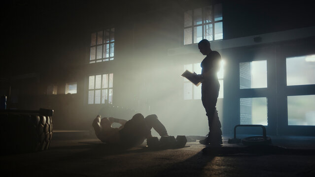 Determined athlete doing sit-ups with personal trainer in dramatic gym setting