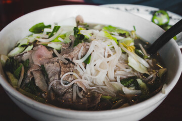 Closeup of a bowl of Pho, a traditional Vietnamese soup. It is traditionally served with a plate of fresh garnishes on the side, including bean sprouts, Thai basil, lime, and chilies