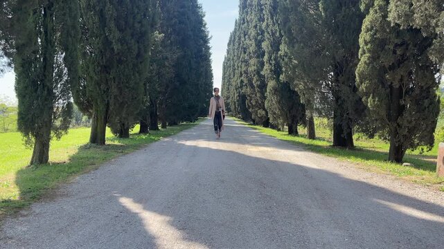 A beautiful woman walking on a gravel road between a row of trees in Tuscany.