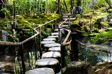 A serene stone path with a bamboo handrail crosses a stream in a lush Japanese garden