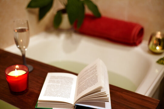 Cozy home spa and relaxation concept with a bathtub filled with fresh flower petals. Wooden bath tray with an open book, scented candles and a glass of wine